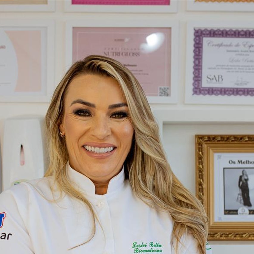 Leslei sorrindo, com jaleco, em frente ao mural de certificações da clínica